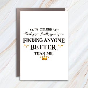 Op de afbeelding: Een witte wenskaart met zwarte tekst: LET'S CELEBRATE the day you finally gave up on FINDING ANYONE BETTER THAN ME. Het ontwerp bevat vier kleine gouden vierpuntige sterren en een gouden kroonpictogram dat onderaan in het midden staat.