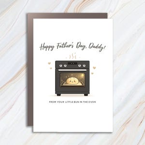 May include: A white greeting card featuring a black oven illustration with a cute, smiling bun inside. The text reads Happy Father's Day, Daddy! at the top and FROM YOUR LITTLE BUN IN THE OVEN at the bottom, accented by small floating hearts.