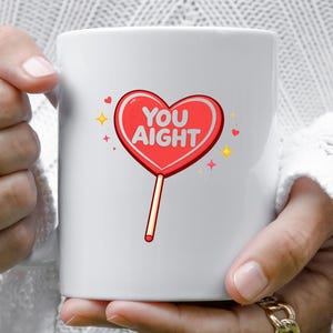 May include: White ceramic mug featuring a red heart-shaped lollipop with the words "YOU AIGHT" in white. The lollipop has a light brown stick and is surrounded by small hearts and starbursts.