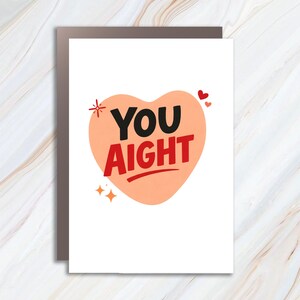 May include: A greeting card with a white background and a peach-colored heart shape. The heart contains the words "YOU AIGHT" in black and red. Small red hearts and starbursts decorate the card.