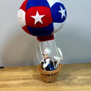 May include: A decorative hot air balloon with a red, white, and blue color scheme. The balloon is made of felt and features white stars. A small basket made of woven wicker holds a figurine wearing a patriotic dress.
