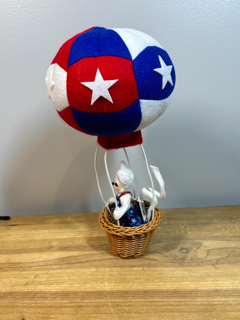 May include: A decorative hot air balloon with a red, white, and blue color scheme. The balloon is made of felt and features white stars. A small basket made of woven wicker holds a figurine wearing a patriotic dress.