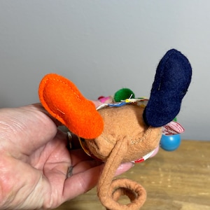 May include: A handmade brown felt toy with orange and navy blue accents. The toy has a curled tail and colorful embellishments. The toy is held in a hand against a wooden surface.