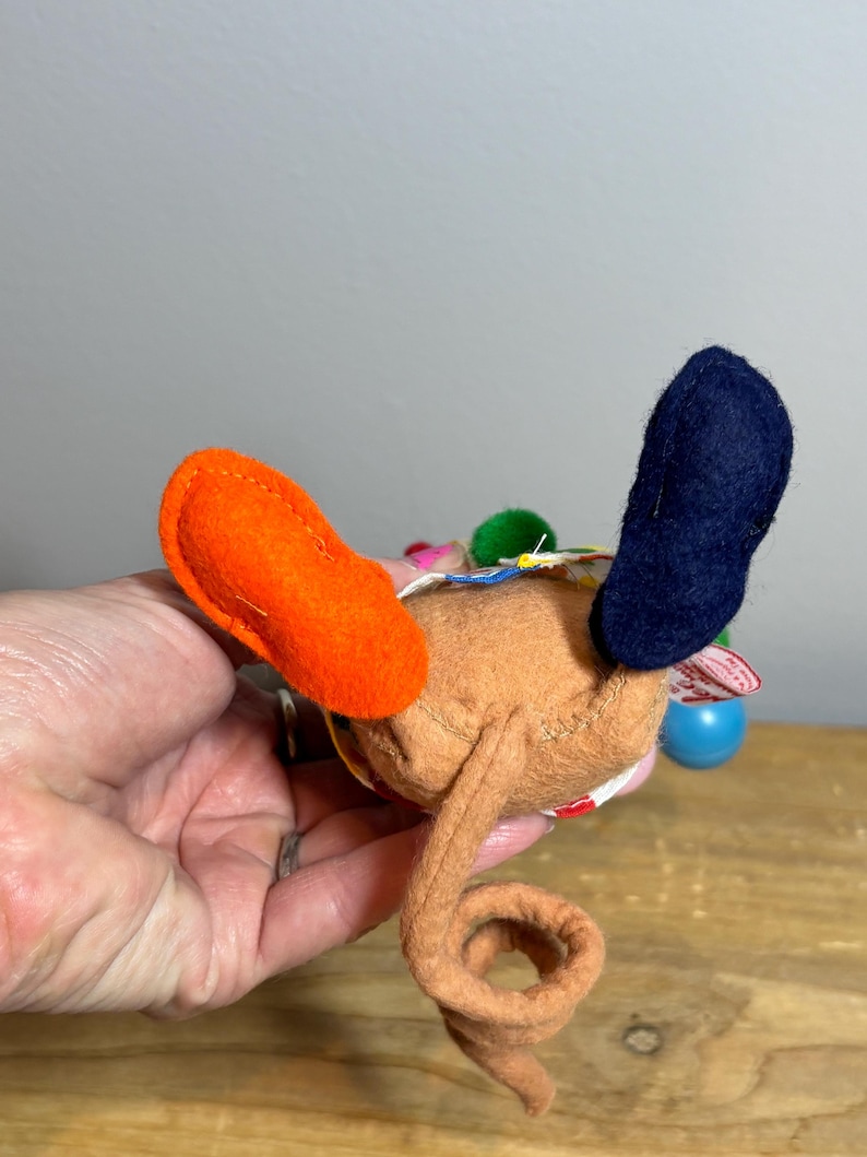 May include: A handmade brown felt toy with orange and navy blue accents. The toy has a curled tail and colorful embellishments. The toy is held in a hand against a wooden surface.