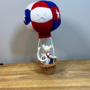 May include: A decorative hot air balloon with a red, white, and blue color scheme. The balloon has white stars and a small woven basket. A white mouse figurine is inside the basket, wearing a red and blue outfit.