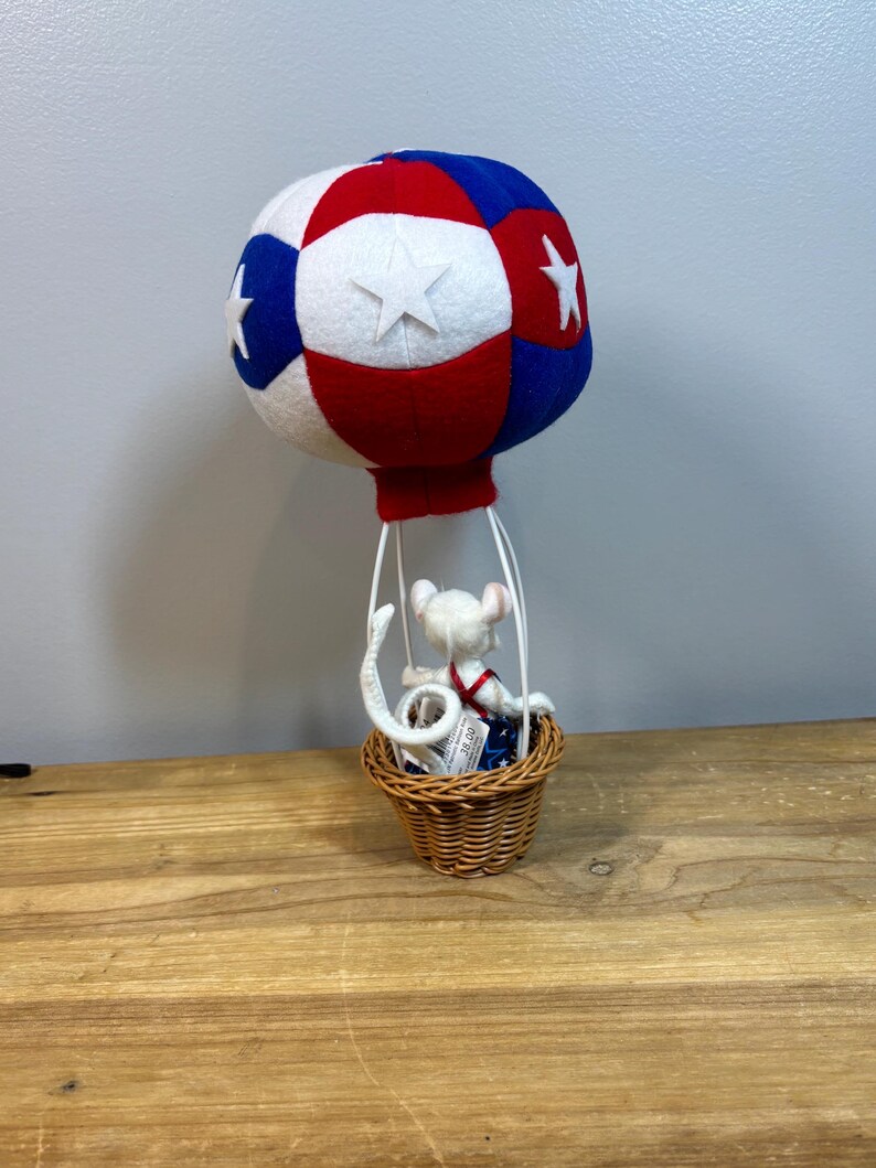 May include: A decorative hot air balloon with a red, white, and blue color scheme. The balloon has white stars and a small woven basket. A white mouse figurine is inside the basket, wearing a red and blue outfit.