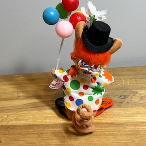 May include: A whimsical clown figurine with orange fur, a black top hat, and a colorful polka-dot outfit. The clown holds a bouquet of colorful balloons. A small brown object is at the clown's feet. The figurine is on a wooden surface.