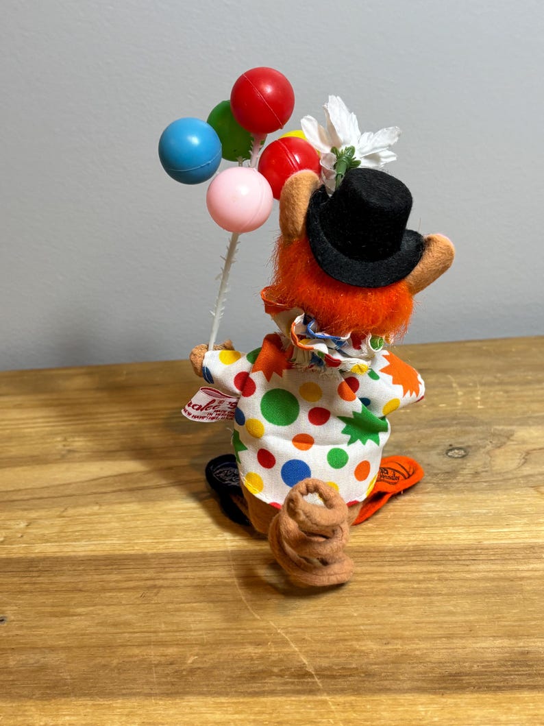 May include: A whimsical clown figurine with orange fur, a black top hat, and a colorful polka-dot outfit. The clown holds a bouquet of colorful balloons. A small brown object is at the clown's feet. The figurine is on a wooden surface.