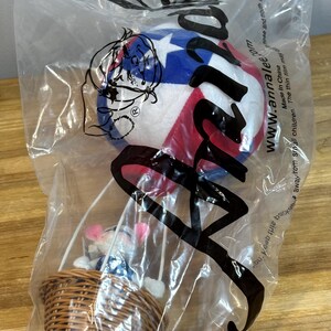 May include: A plush hot air balloon toy in a clear plastic bag. The balloon is white, red, and blue with a star pattern. A woven basket holds small plush figures. The bag has the text "www.annalee.com".