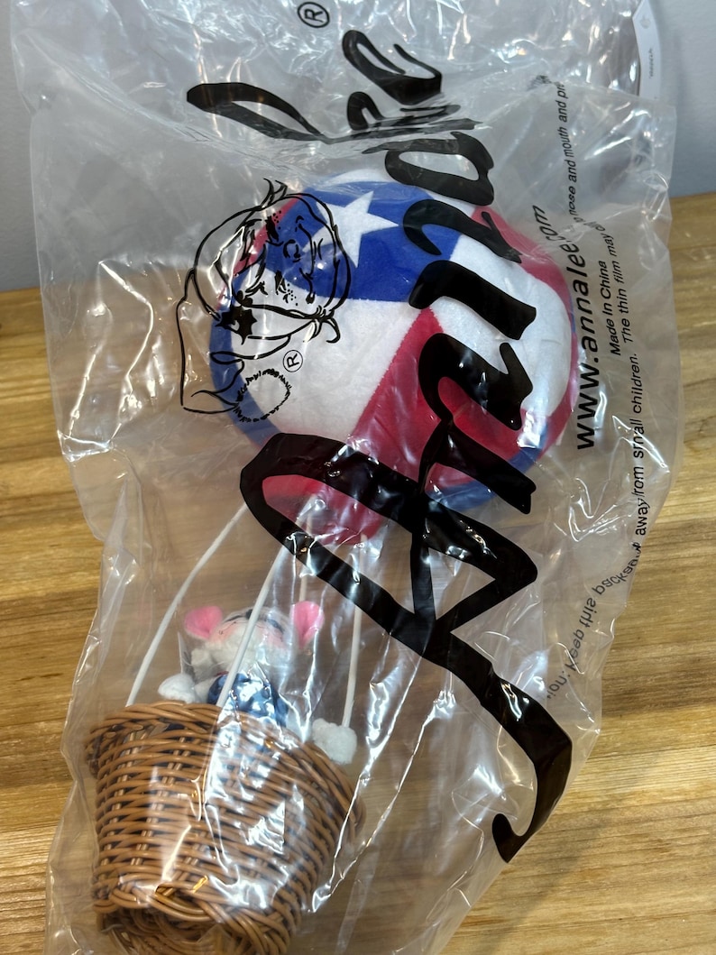 May include: A plush hot air balloon toy in a clear plastic bag. The balloon is white, red, and blue with a star pattern. A woven basket holds small plush figures. The bag has the text "www.annalee.com".