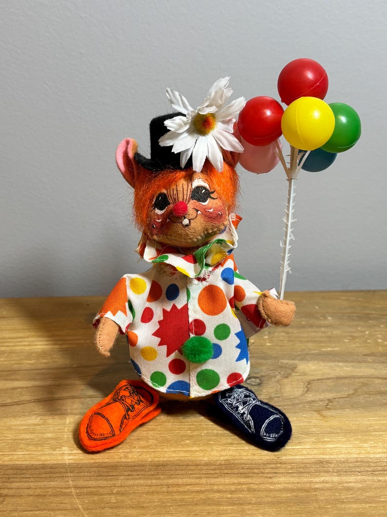 May include: A whimsical clown doll with orange fur, a black top hat, and a daisy. The doll wears a colorful polka-dot shirt and holds a bunch of balloons. The clown has orange and navy blue shoes. A vintage-style collectible.