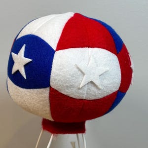 May include: A patriotic red, white, and blue decorative hot air balloon with star accents. The balloon is made of felt and has a red base. The balloon is supported by a white wire frame, with a small figure inside.