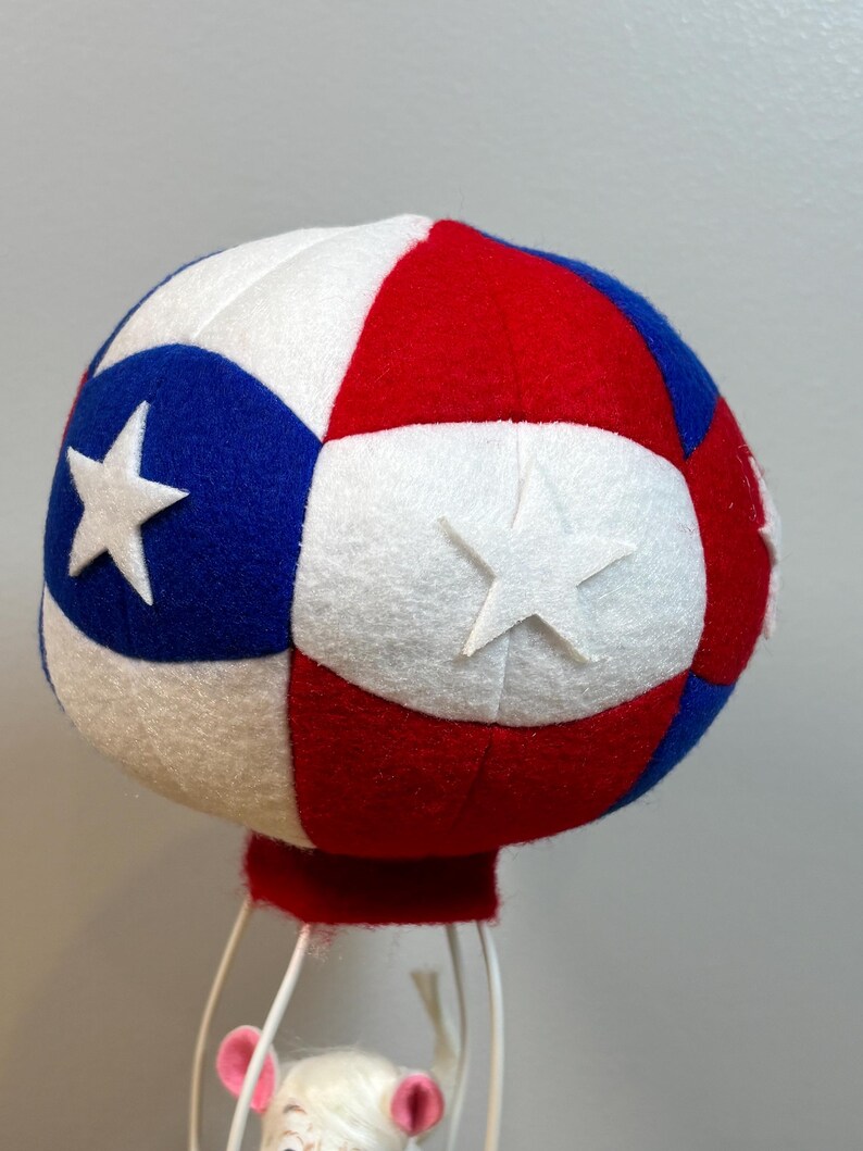 May include: A patriotic red, white, and blue decorative hot air balloon with star accents. The balloon is made of felt and has a red base. The balloon is supported by a white wire frame, with a small figure inside.