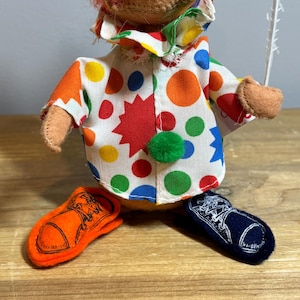 May include: A vintage clown doll with orange hair, a red nose, and a colorful polka-dot outfit. The doll has orange and navy blue shoes. The clown's outfit features a white shirt with large red, yellow, green, and blue polka dots.