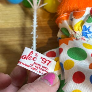 May include: A vintage clown doll with orange hair, a black hat, and a colorful polka-dot outfit. The doll holds a cluster of miniature balloons. A tag reads "alee '97 MADE ONLY IN MEREDITH, NEW HAMPSHIRE, U.S."