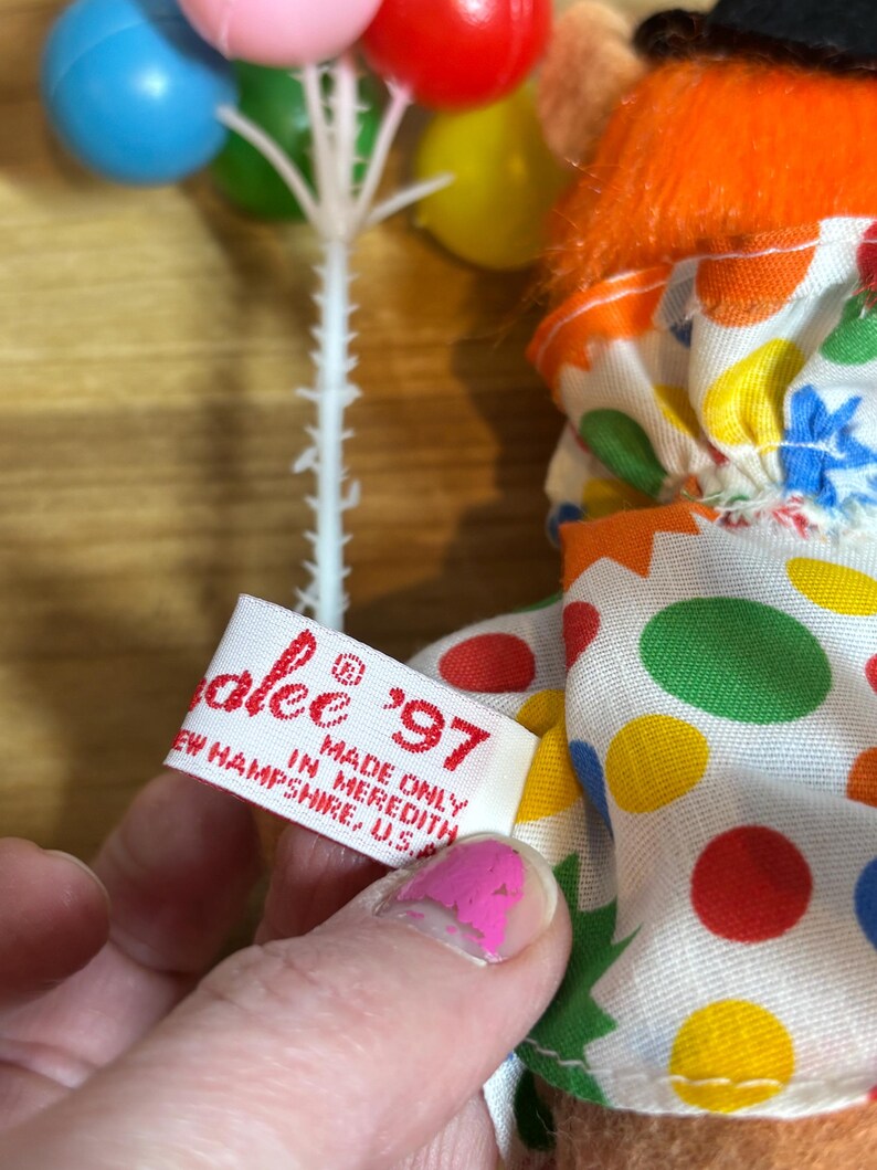 May include: A vintage clown doll with orange hair, a black hat, and a colorful polka-dot outfit. The doll holds a cluster of miniature balloons. A tag reads "alee '97 MADE ONLY IN MEREDITH, NEW HAMPSHIRE, U.S."