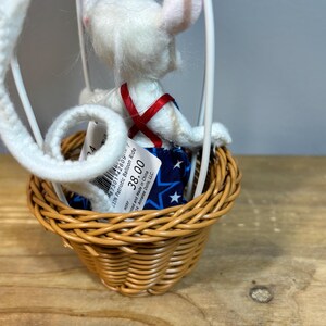 May include: A white mouse doll in a patriotic balloon ride. The mouse wears a blue star-patterned outfit with a red cross-strap. The balloon basket is woven, and the frame is white. The doll is a collectible item.