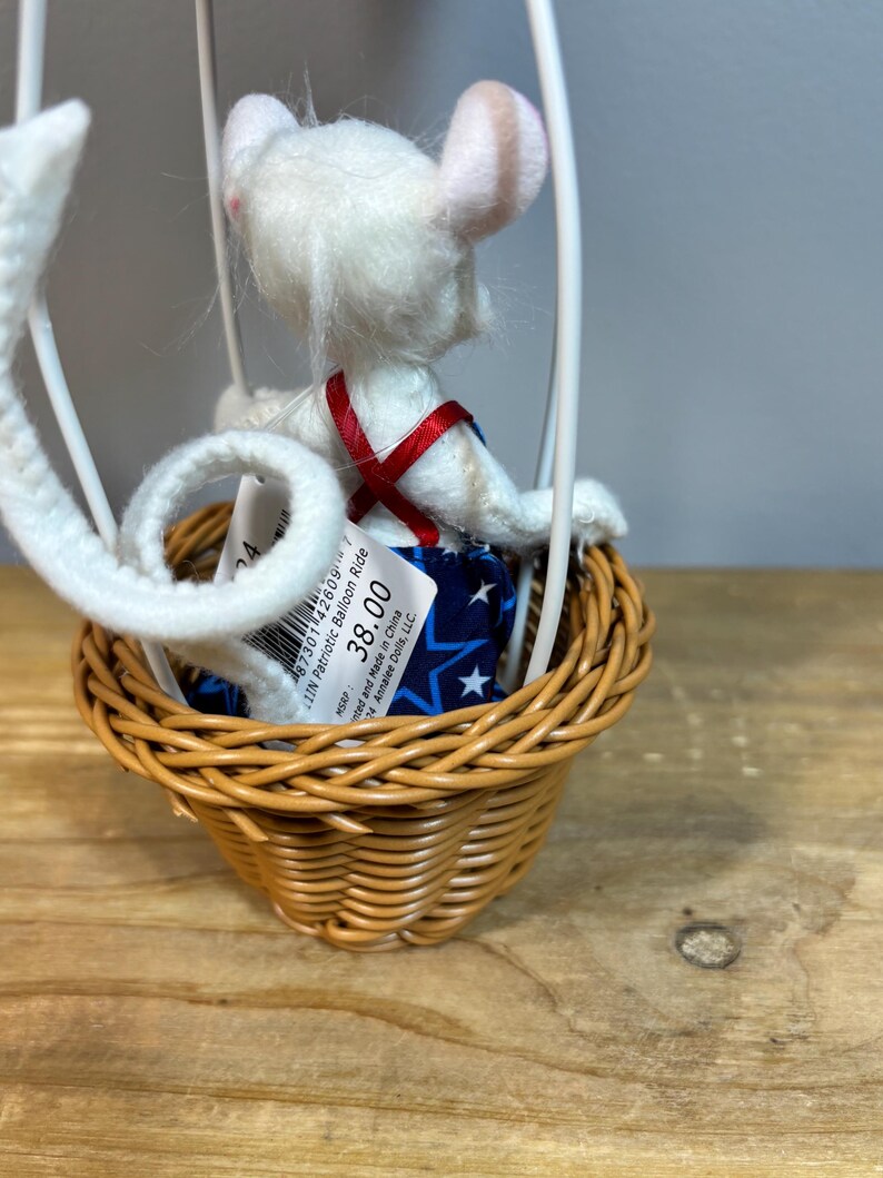 May include: A white mouse doll in a patriotic balloon ride. The mouse wears a blue star-patterned outfit with a red cross-strap. The balloon basket is woven, and the frame is white. The doll is a collectible item.