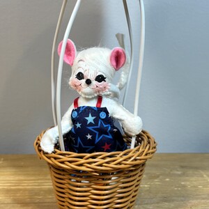 May include: A whimsical mouse doll in a hot air balloon. The white mouse has pink ears and is wearing a blue overall with white stars. The balloon basket is woven, and the balloon top is red. The balloon frame is white.