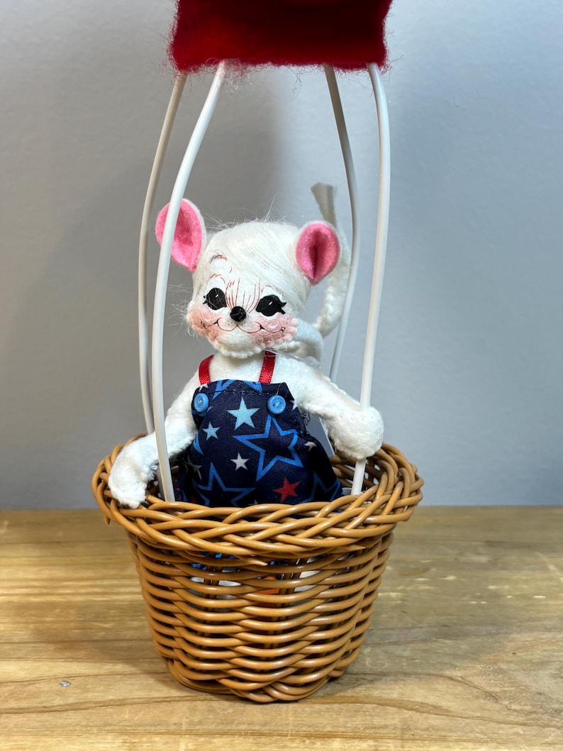 May include: A whimsical mouse doll in a hot air balloon. The white mouse has pink ears and is wearing a blue overall with white stars. The balloon basket is woven, and the balloon top is red. The balloon frame is white.