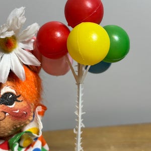 May include: A vintage clown doll with orange hair and a daisy flower. The clown is holding a bouquet of colorful balloons: red, yellow, green, pink, and blue. The clown is wearing a polka dot outfit.