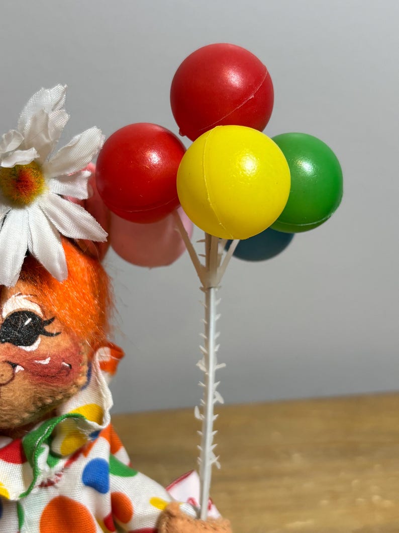 May include: A vintage clown doll with orange hair and a daisy flower. The clown is holding a bouquet of colorful balloons: red, yellow, green, pink, and blue. The clown is wearing a polka dot outfit.