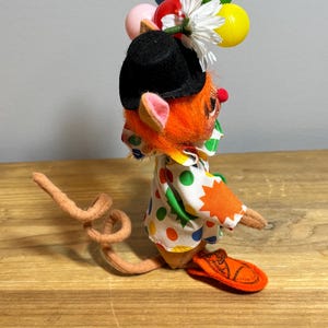 May include: A whimsical clown mouse figurine with orange fur, a black hat, and a polka-dot shirt. The mouse holds colorful balloons and a white flower. The figurine has a long, curled tail and orange sandals.