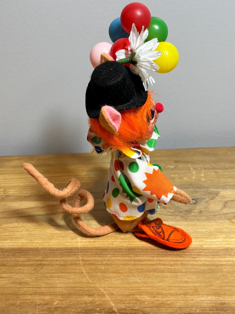 May include: A whimsical clown mouse figurine with orange fur, a black hat, and a polka-dot shirt. The mouse holds colorful balloons and a white flower. The figurine has a long, curled tail and orange sandals.