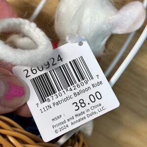 May include: A white plush toy in a wicker basket, part of a patriotic balloon ride set. The toy has a tag that reads "11IN Patriotic Balloon Ride". The tag also includes a barcode and the text "Printed and Made in China".