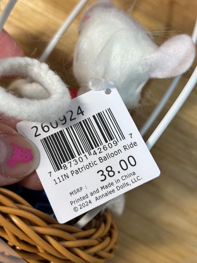 May include: A white plush toy in a wicker basket, part of a patriotic balloon ride set. The toy has a tag that reads "11IN Patriotic Balloon Ride". The tag also includes a barcode and the text "Printed and Made in China".
