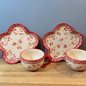 May include: A set of two floral-patterned plates and two matching teacups. The plates are shaped like four-leaf clovers with a red scalloped edge. The teacups have red handles and a similar floral design. The background is a blue wall and a wooden surface.