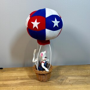 May include: A whimsical, handmade hot air balloon with a red, white, and blue color scheme. The balloon features white stars and is attached to a woven basket. A small mouse figurine wearing overalls is in the basket.