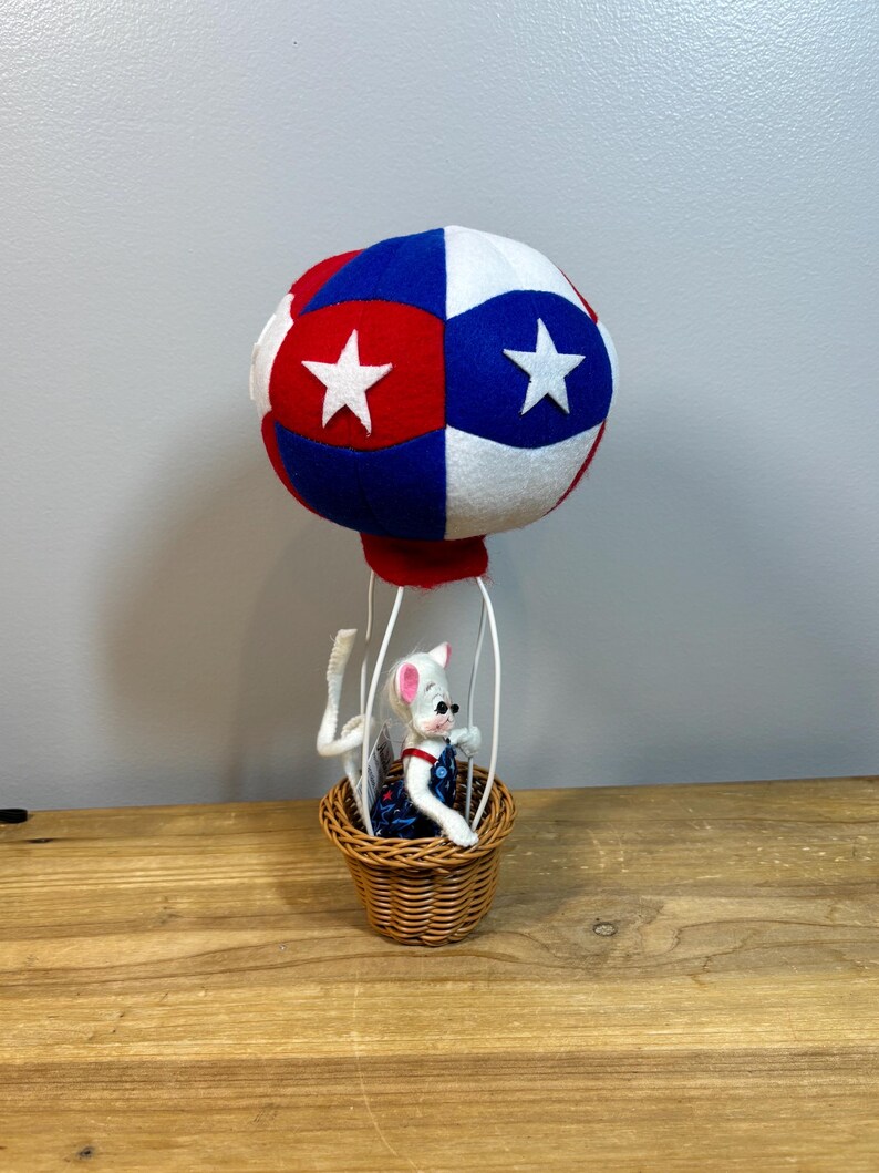 May include: A whimsical, handmade hot air balloon with a red, white, and blue color scheme. The balloon features white stars and is attached to a woven basket. A small mouse figurine wearing overalls is in the basket.
