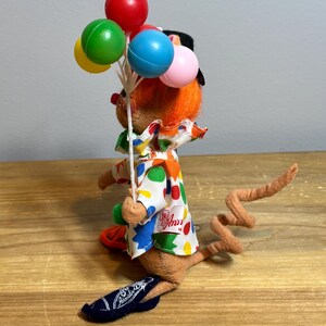 May include: A vintage Annalee clown mouse doll holding a bouquet of colorful balloons. The doll has orange hair, a black hat, and a white shirt with colorful polka dots. The mouse is wearing a blue shoe with the Annalee logo.