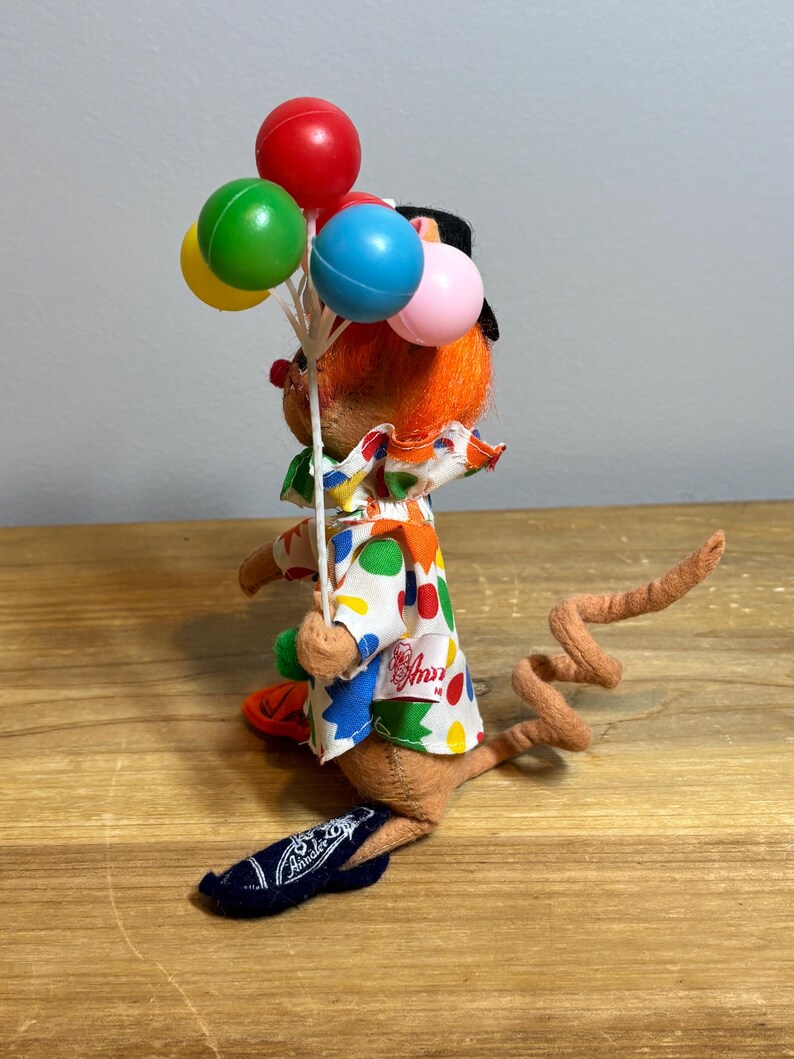 May include: A vintage Annalee clown mouse doll holding a bouquet of colorful balloons. The doll has orange hair, a black hat, and a white shirt with colorful polka dots. The mouse is wearing a blue shoe with the Annalee logo.