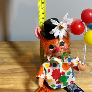 May include: A vintage clown doll with orange hair, a black top hat, and a white flower. The doll wears a colorful polka-dot shirt and holds a cluster of red, blue, and yellow balloons. The doll is approximately 7 inches tall.