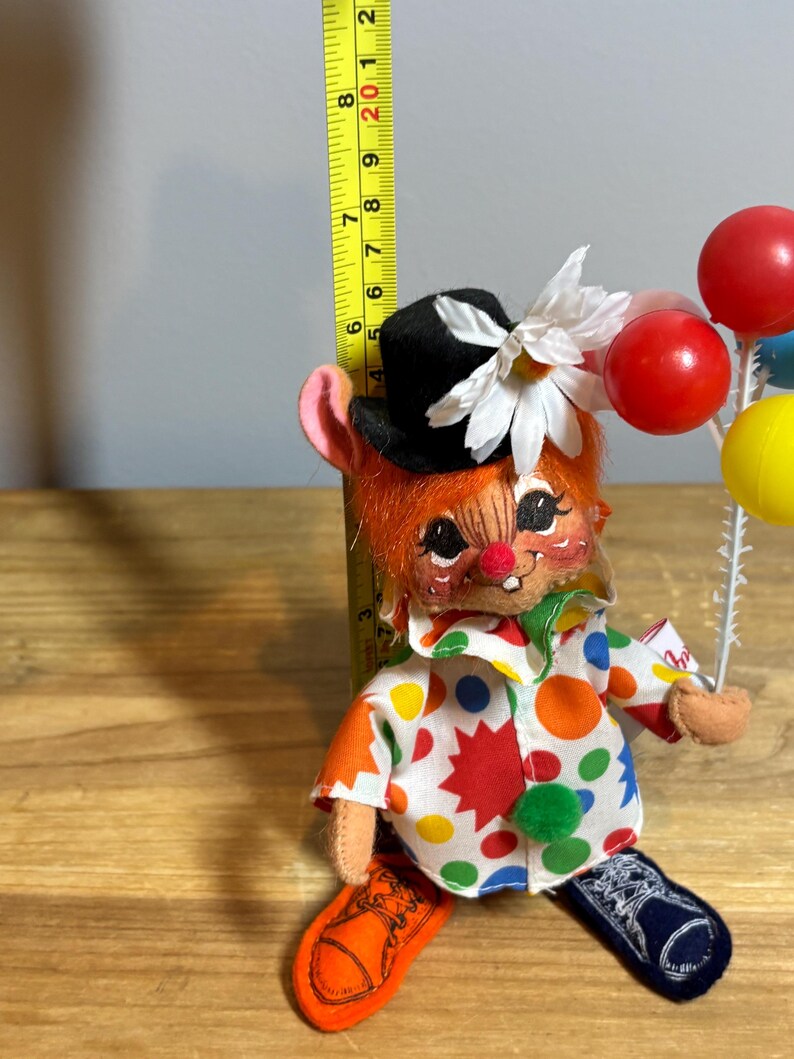 May include: A vintage clown doll with orange hair, a black top hat, and a white flower. The doll wears a colorful polka-dot shirt and holds a cluster of red, blue, and yellow balloons. The doll is approximately 7 inches tall.