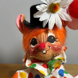 May include: A vintage clown doll with orange hair, a black top hat, and a white daisy. The doll has a red nose, rosy cheeks, and a colorful polka-dot shirt. It is holding a red balloon.