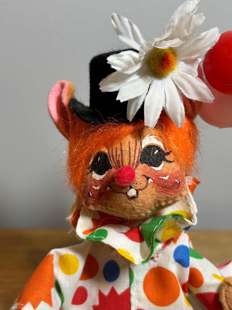 May include: A vintage clown doll with orange hair, a black top hat, and a white daisy. The doll has a red nose, rosy cheeks, and a colorful polka-dot shirt. It is holding a red balloon.