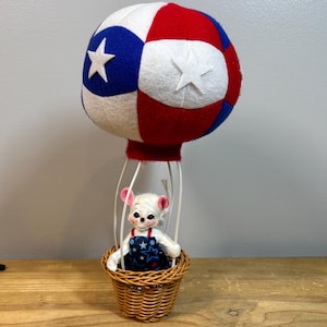 May include: A whimsical hot air balloon decoration featuring a white mouse in a woven basket. The balloon is red, white, and blue with star accents. The mouse wears a blue and white star-patterned outfit. The balloon is supported by a wire frame.