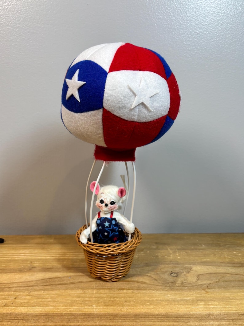 May include: A whimsical hot air balloon decoration featuring a white mouse in a woven basket. The balloon is red, white, and blue with star accents. The mouse wears a blue and white star-patterned outfit. The balloon is supported by a wire frame.