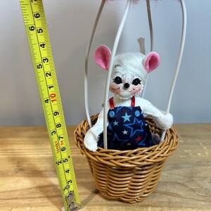 May include: A whimsical handmade toy mouse in a hot air balloon. The mouse has white fur, pink ears, and is wearing blue overalls with a star pattern. The balloon is red and blue, and the basket is woven. The toy is approximately 6 inches tall.