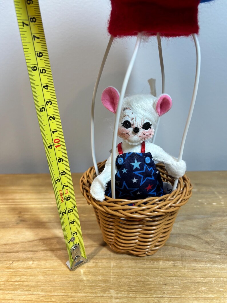 May include: A whimsical handmade toy mouse in a hot air balloon. The mouse has white fur, pink ears, and is wearing blue overalls with a star pattern. The balloon is red and blue, and the basket is woven. The toy is approximately 6 inches tall.