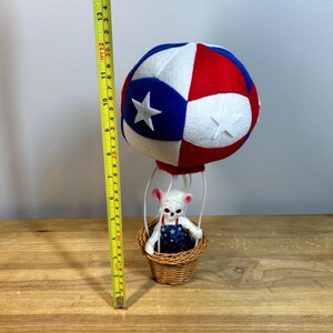 May include: A patriotic-themed hot air balloon decoration. The balloon is red, white, and blue with star accents. A small woven basket holds a white mouse figurine wearing a blue and white star-patterned outfit. The balloon measures approximately 14 inches in height.
