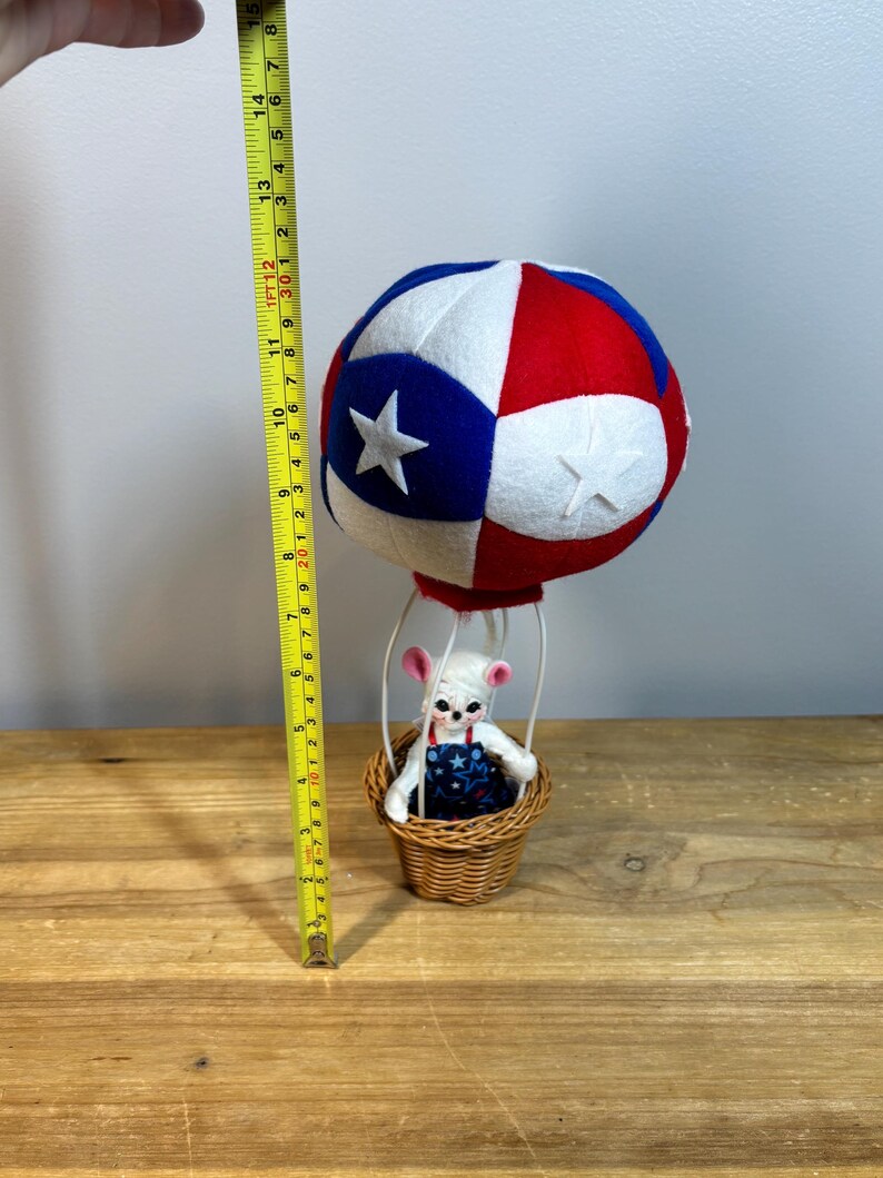 May include: A patriotic-themed hot air balloon decoration. The balloon is red, white, and blue with star accents. A small woven basket holds a white mouse figurine wearing a blue and white star-patterned outfit. The balloon measures approximately 14 inches in height.