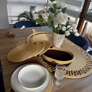 May include: A dining table set with woven placemats, coasters, and a basket. White plates with gold rims are stacked with a bowl. A vase of white roses and tulips provides a floral centrepiece.