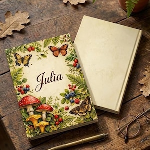 Può includere: Un diario personalizzato con il nome "Julia" sulla copertina, circondato da illustrazioni di farfalle, funghi e bacche. Un diario vuoto è accanto su una superficie di legno.