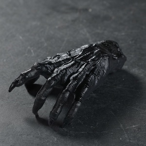 May include: A black, sculpted hand figurine featuring elongated, claw-like fingers and a textured, wrinkled surface. The hand is positioned in a reaching gesture with a defined wrist area, showcasing detailed anatomical ridges and deep grooves.