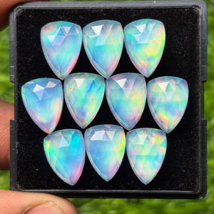 May include: A collection of ten pear-shaped opals, each with a faceted surface. The opals display a play of color, with iridescent hues of blue, green, and pink. They are arranged in a black tray.