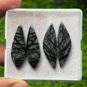K&ouml;nnte beinhalten: Vier graue und schwarze Edelstein-Cabochons in einer wei&szlig;en Schale. Die tropfenf&ouml;rmigen Steine weisen ein Netzwerk aus schwarzen Linien auf grauem Hintergrund auf. Diese Cabochons sind wahrscheinlich f&uuml;r die Schmuckherstellung oder andere Bastelarbeiten bestimmt.